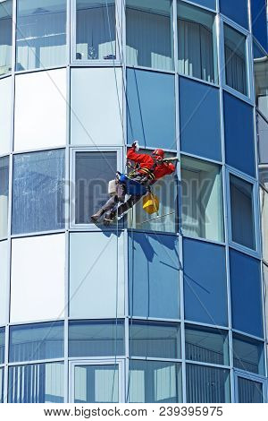 Worker In A Helmet Washes The Window Of A High-rise Building From The Outside, Hanging On A Rope