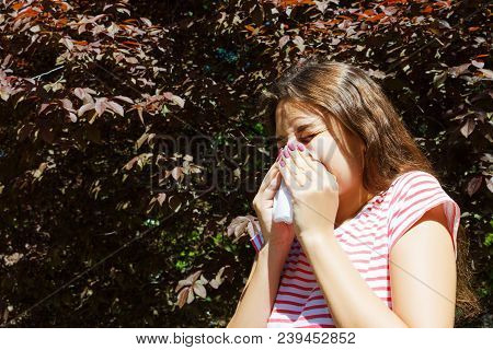 Sneezing Young Girl With Napkin Among Blooming Trees In Park. Allergy Concept