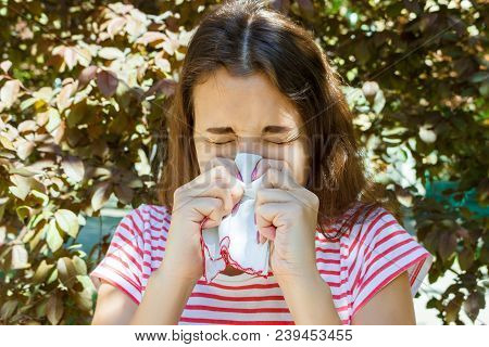 Sneezing Young Girl With Napkin Among Blooming Trees In Park. Allergy Concept