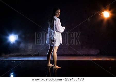 Girl In White Shirt Stands In Small Pool During Photoshoot With Water And Black Wall Illuminated Col