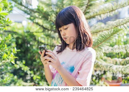 Little Cute Asian Kid Girl In Pink T-shirt Looking At Device And Using Wireless Smartphone Send Mess