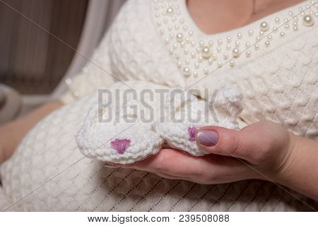 White Handmade Knited Booties In Shape Of Rabbits In Hand Of Pregnant Woman Dressed In Knitted White