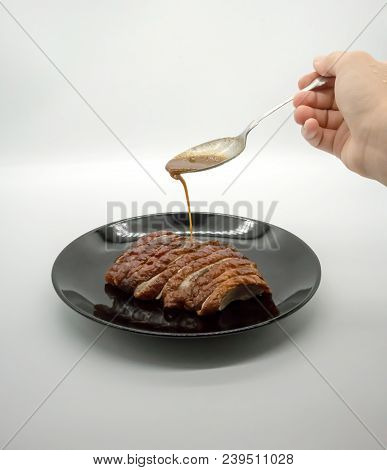 Woman Hand Holding Silver Spoon Adding Sauce On Top Of Roasted Duck On Black Ceramic Dish Isolated O