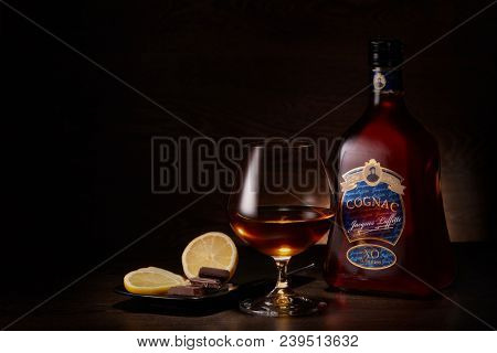 Bottle of cognac Jacques Laffitte XO Superior, glass with cognac and plate with pieces of lemon and chocolate stands on a table on a dark wooden background.