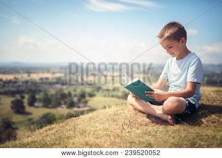 Boy sitting and relaxing on a hill reading a book in a meadow concept for education and relaxing