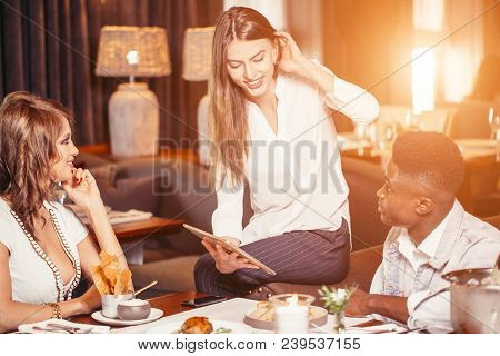 Happy Smiling Young Mates In Cafe. Young Woman Sitting At Cafe Presents Her Friends New Digital Tabl