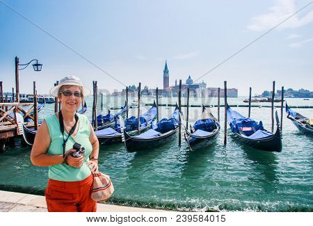 Mature Tourist Woman Travelling In Italy Stays Against Row Of Gondolas Moored By Saint Mark Square A