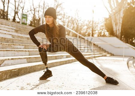 Image of disabled athletic girl in sportswear doing sit ups and stretching bionic leg at stairs outdoors