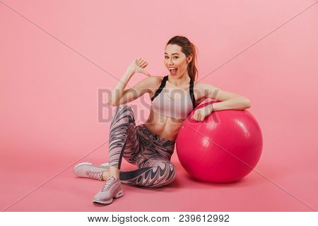 Pleased sportswoman sitting on the flloor with fitness ball while indicates itself and looking at the camera over pink background
