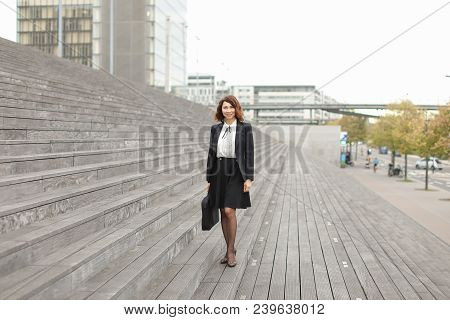 American Businesswoman Standing On Stairs With Bag And High Buildings In Background. Concept Of Prof