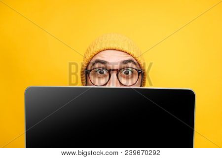 Young Expressive Hipster In Hat And Eyeglasses Covering Face With Laptop On Yellow.