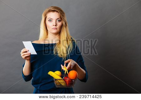 Worried Woman Holding Shopping Basket With Fruits Looking At Bill Receipt Being Scared Of Huge Price