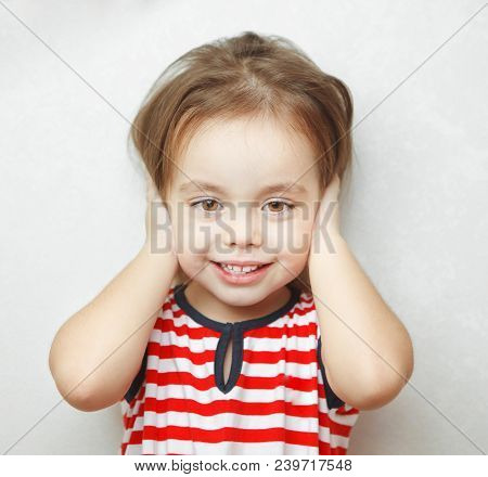 Adorable Little Girl With Sincere Smile And Big Brown Eyes Covers Her Ears With Palms Dressed In Str