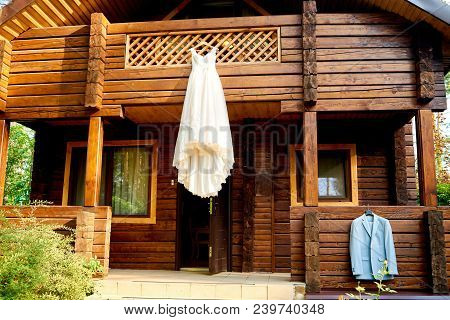 Beautiful White Wedding Dress And Groom Suit Hanging On Balcony Of Brown Wooden House Outdoors, Copy