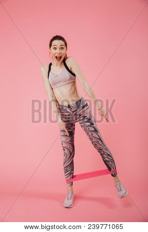 Full length image of Surprised happy sportswoman doing fitness exrcise and pointing down while looking at the camera over pink background