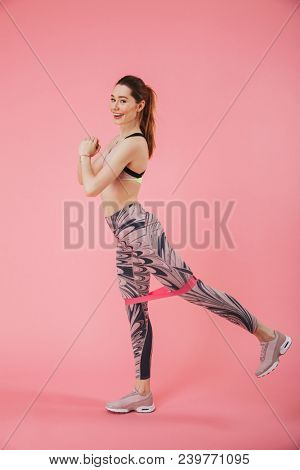 Vertical side view image of happy sportswoman doing fitness exrcise and looking at the camera over pink background