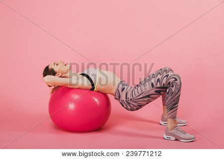 Side view of Funny sportswoman doing exrcise with fitness ball and looking up over pink background