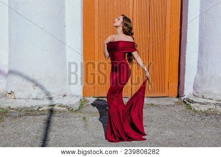 Beautiful Young Woman In A Long Red Evening Gown With A Train Standing Next To The Old Door