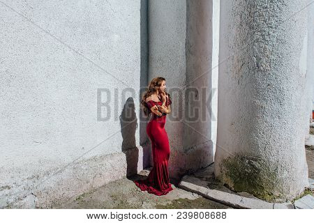 Beautiful Young Woman In A Long Red Evening Gown With A Train Standing Next To The Old Wall