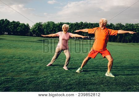 Senior Man And Woman Are Outdoors In A Park. They Are Wearing Casual Exercise Clothing And Practicin