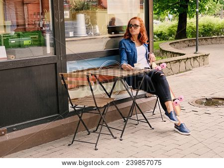 Full Body Imgae Of Redhead Female Drinks Coffee At The Table In A Cafe On A Street.