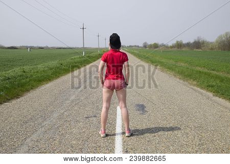 A Young Woman In A Black Cap And Red Clothes Is Standing In The Middle Of The Road.