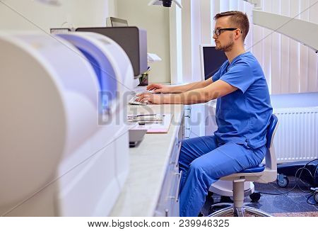 Handsome Male Dentis Using Computer In A Room With Medical Equipment On Background.