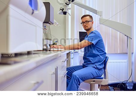 Handsome Male Dentis Using Computer In A Room With Medical Equipment On Background.