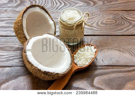 Two Halves Of Coconut With Shavings In A Glass Jar And A Wooden Spoon On A Wooden Table