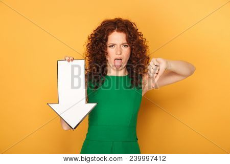 Portrait of a disppointed redhead woman in dress pointing down with an arrow isolated over yellow background
