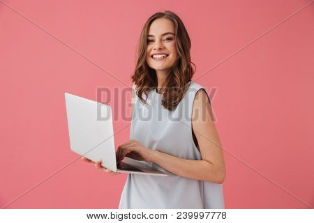 Image of cheerful young woman standing isolated over pink background using laptop computer. Looking camera.