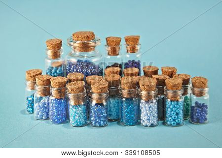 Purple And Cyan Beads In Glass Jars On Bright Blue Background. Beads In A Transparent Container With