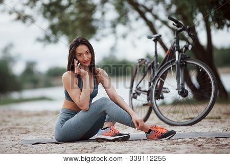 Making A Call. Female Cyclist With Good Body Shape Sitting Near Her Bike On Beach At Daytime.