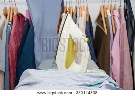 Iron Stands On The Ironing Board On The Background Of A Lot Of Things And Clothes. Have To Iron A Lo