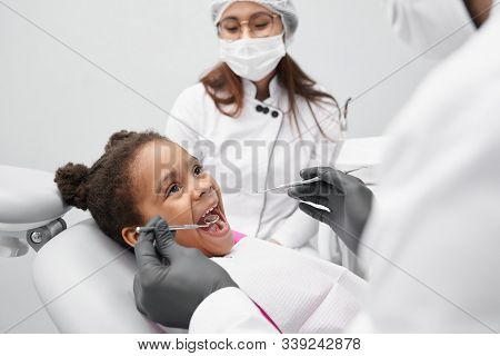 View From Side Of Funny Girl Lying On Dentist Chair With Open Mouth While Doctor Examining Teeth. Pr