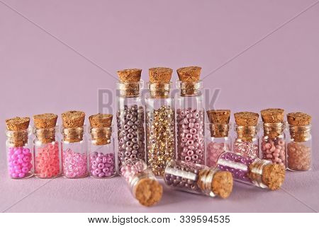 Beige, Pink And Yellow Beads In Glass Jars On A Bright Purple Background. Beads In A Transparent Con