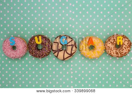 Group Of Delicious Colorful Glazed Donuts Near Green Background With White Hearts Pattern And The Wo