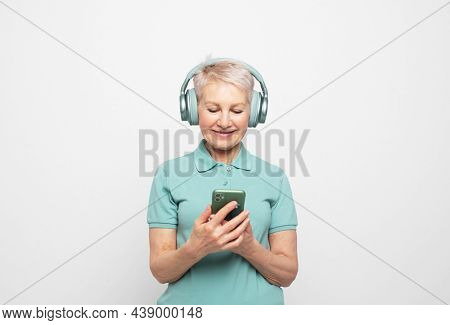 Happy mature short haired woman smiling broadly posing against whte grey background in wireless headphones, jolding modern smartphone, enjoying clean high quality sound, listening to music online