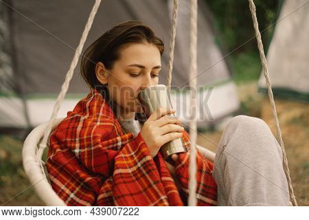 Beautiful Girl Wrapped In Red Plaid Drinking Tea In A Cozy Hanging Chair Outdoors. Girl Holding Mug 