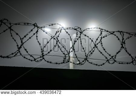 Silhouette Of Barbed Wire On A Fence Against A Background Of Blurry Lamp Posts At Foggy Night.