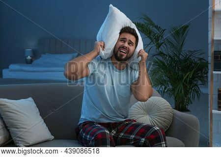 Unhappy Young Man Covering Ears With Pillow In Living Room At Night. Noisy Neighbours