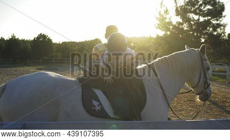 A Small Jockey Boy In A Helmet Climbs A White Horse. Little Boy Learns With An Instructor Horse Ridi
