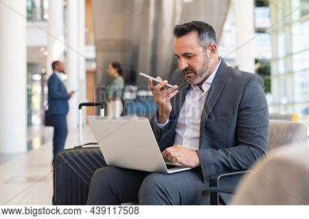 Serious businessman sitting on couch in airport lounge using laptop and sending voice message with smartphone. Mature business man working on computer while using voice command recorder on phone.