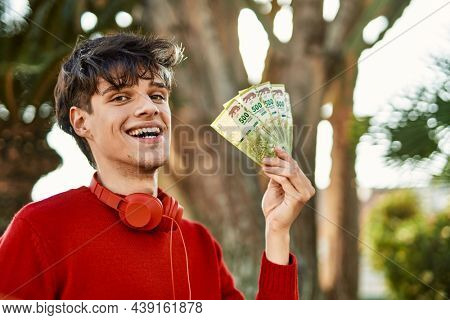 Young hispanic man using headphones holding argentinian pesos at the city