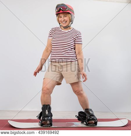Elderly Woman Imitates Snowboarding In The Studio On A White Background.