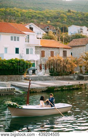 Man Rolls A Woman On A Boat Decorated With Flowers Against The Background Of The Pier And Old Houses