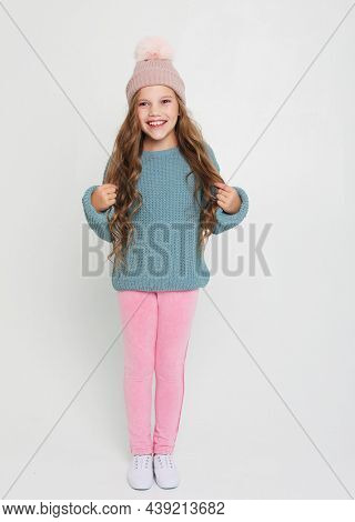 emotion, childhood and people concept: funny child girl with curly haor dressed blue sweater and winter hat jumping on white background