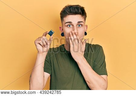 Young caucasian boy with ears dilation holding sdxc card covering mouth with hand, shocked and afraid for mistake. surprised expression 