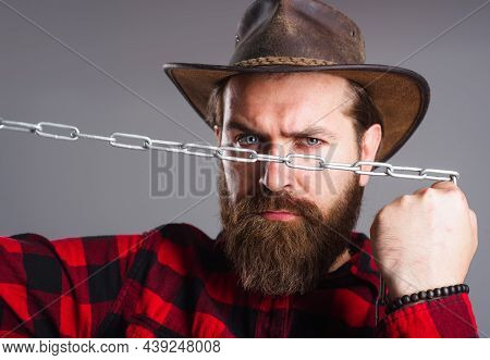Brutal Man In Cowboy Hat With Chain. Serious Bearded Male In Plaid Shirt Trying To Break Metal Chain