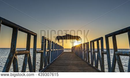 Sunrise Over The Red Sea. A Wooden Path With A Metal Railing Goes Over The Water To The Canopy.  The